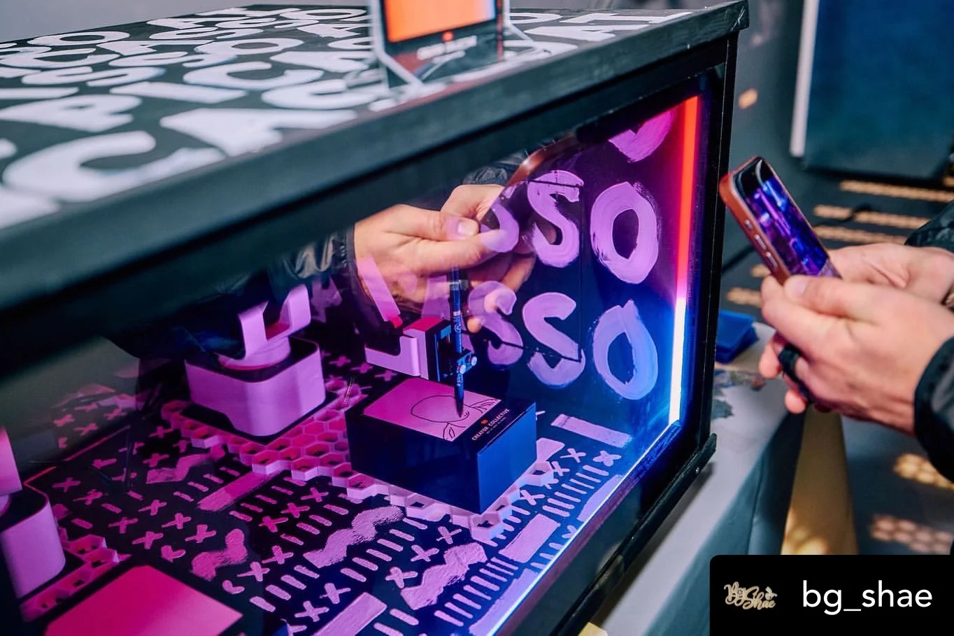 Close view of a hand retrieving artwork inside the illuminated SketchBot, beside a person holding a phone
